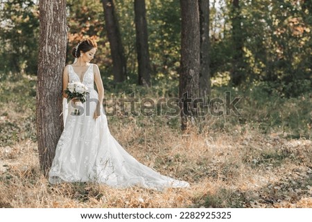 Wedding photo in nature. The bride is standing near a tree in a beautiful dress, holding her bouquet of white roses, looking to the side. Beautiful makeup of the bride. Portrait.