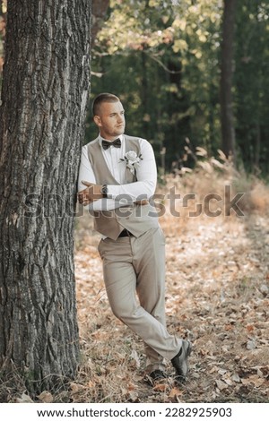 Portrait of the groom. The groom in a light gray suit is standing in the forest, leaning against a tree and looking away. Wedding in nature