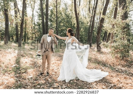 Wedding photo. The bride and groom are walking in the forest. The groom holds a bouquet and kisses the hand of his beloved, and she beautifully throws up her long veil. Couple in love. Summer light.