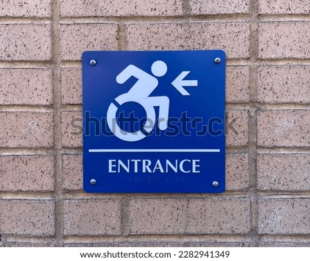 Close-up of blue wheelchair accessibility sign with forward-leaning head and motioning arms next to word "Entrance" and arrow pointing left screwed to brick wall on the outside of building in NYC
