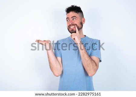 Positive Young caucasian man wearing blue t-shirt over white background advert promo touch finger teeth