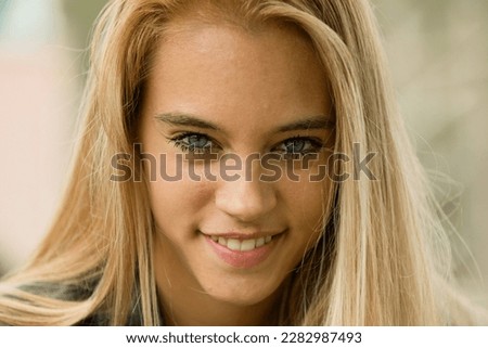 Blue eyes of a gorgeous young blonde woman, outdoors in daylight, with a blurred background, amidst nature and greenery, among the leaves of a public park or a forest. 