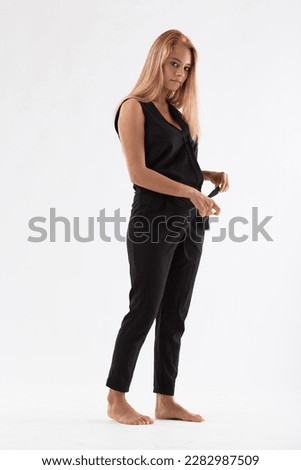 Barefoot blonde girl wearing a black jumpsuit, striking fashion poses, walking, fastening, and changing looks. Her bare feet contrast both the white background and the black outfit. She has blue eyes 