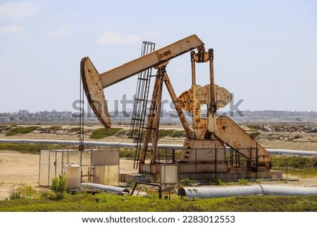 a rusty metal oil rig surrounded by dirt and green plants and grass with clear blue sky at Bolsa Chica Ecological Reserve in Huntington Beach California USA