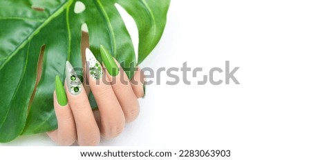 Female hand with vacation stiletto nail design. Glitter green nail polish manicure with rhinestones and glitter nail art. Female model hand hold tropic leaf on white background. Copy space Royalty-Free Stock Photo #2283063903