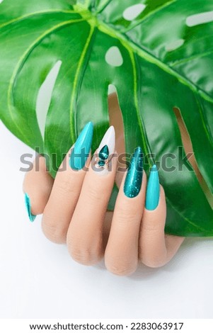 Female hand with vacation stiletto nail design. Glitter cyan nail polish manicure with rhinestones spider nail art. Female model hand hold tropic leaf on white background. Copy space. Place for text. Royalty-Free Stock Photo #2283063917