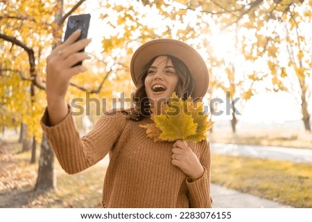 Attractive young smiling woman walking in autumn park taking selfie pictures using smartphone, holding yellow leaves in her hands, having happy mood, fashion style trend. Copy space