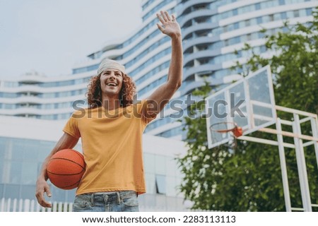 Bottom view young man 20s yellow t-shirt bandana waving hand say hello playing basketball hold ball on playground in free time rest relax in city outdoors on open air. Urban lifestyle leisure concept
