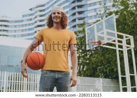 Young happy man 20s wearing yellow t-shirt bandana hold ball look aside playing basketball on playground in free time walking rest relax in city outdoors on open air. Urban lifestyle leisure concept