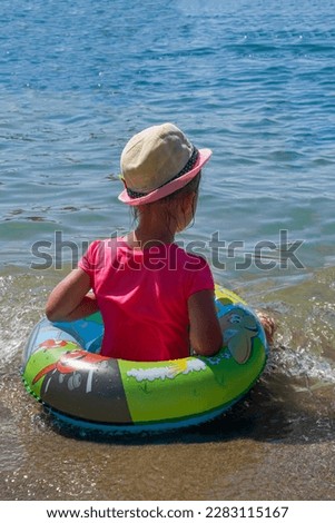 A girl in a circle on the beach, rear view. A child in a pink T-shirt and straw hat sits on the seashore in the water and looks at the waves. Sea vacation concept. Summer mood.