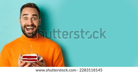 Close-up of happy adult man celebrating birthday, holding bday cake with candle and making wish, standing against turquoise background.