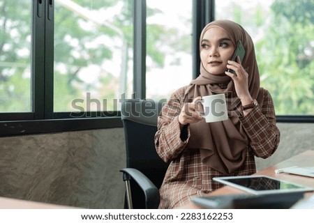  Asian Muslim woman wearing hijab holding a cup of coffee while talking on a mobile phone in her office.