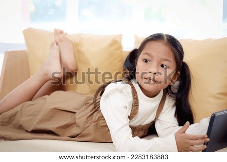 Asian girl using digital tablet while lying down on yellow sofa in living room at home, happy little kid using hi-tech devices to reading book online, child enjoy resting and watching movie on holiday