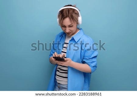 close-up photo of a bright smiling blond young woman dressed in a striped sweater and casual shirt with white wireless headphones communicates in the application in a smartphone on a blue background