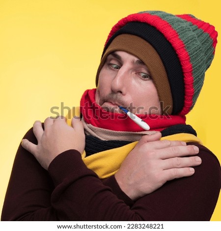 A young man in warm clothes on a yellow background holds an electronic thermometer in his mouth and is shocked by the high temperature Royalty-Free Stock Photo #2283248221