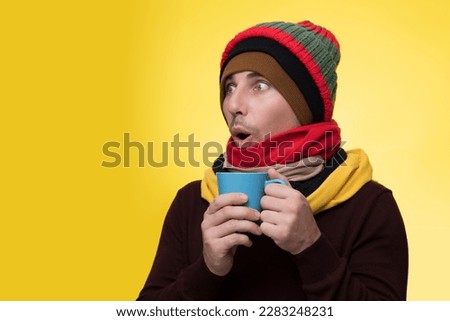 A young man in warm clothes, several hats on a yellow background holds a cup of tea and warms himself during a cold Royalty-Free Stock Photo #2283248231