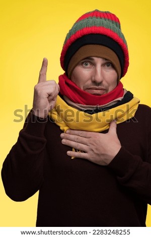 A man on a yellow background in several hats and scarves holds an index finger, attention cold season is near. Royalty-Free Stock Photo #2283248255