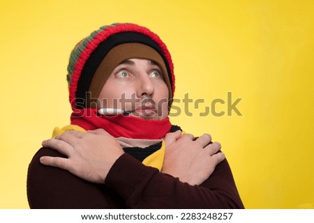 A young man in warm clothes on a yellow background holds an electronic thermometer in his mouth and is shocked by the high temperature Royalty-Free Stock Photo #2283248257