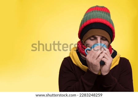 A young man in warm clothes, several hats on a yellow background holds a cup of tea and warms himself during a cold Royalty-Free Stock Photo #2283248259