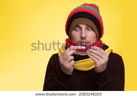 A young man in warm clothes on a yellow background holds an electronic thermometer in his mouth and is shocked by the high temperature Royalty-Free Stock Photo #2283248265