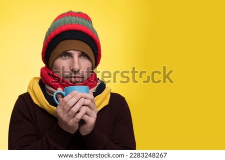 A young man in warm clothes, several hats on a yellow background holds a cup of tea and warms himself during a cold Royalty-Free Stock Photo #2283248267