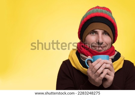 A young man in warm clothes, several hats on a yellow background holds a cup of tea and warms himself during a cold Royalty-Free Stock Photo #2283248275