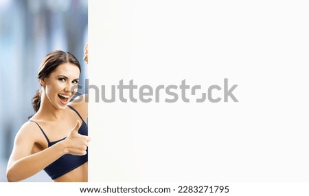 Image of excited brunette woman in fitness wear show thumb up, signboard with copy space area. Fit girl with happy wide open mouth, peek out mock up white placard, blurred modern gym center.