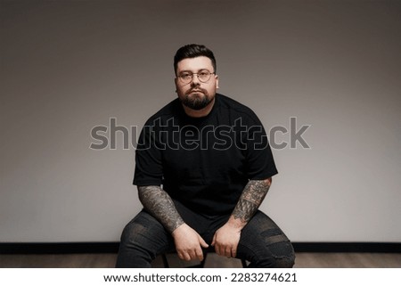 Content young plus size bearded male model in black outfit and eyeglasses with tattooed arms sitting on chair against gray background in studio Royalty-Free Stock Photo #2283274621