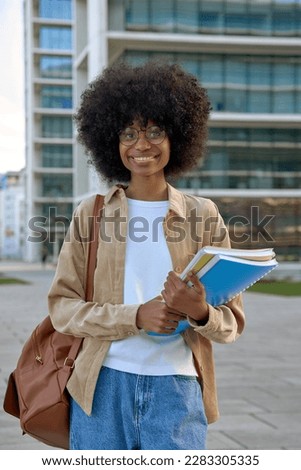 A vertical portrait of African American girl with curly afro hairstyle stands with backpack and holds exercise books in university campus. Smart student look at camera on city background, copy space.