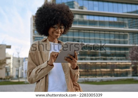 A young woman student using touchscreen of pc tablet standing in university campus. An African American cheerful female business freelancer entrepreneur using app project for online work outdoors.