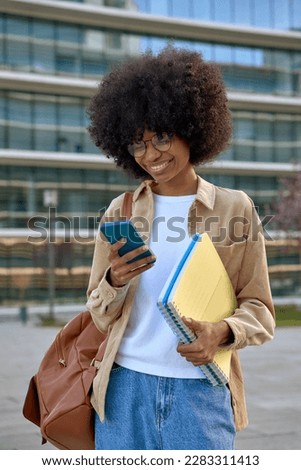 Vertical portrait of happy African American student girl enjoy online communication, hold exercise books with a backpack, scroll social media app on cell phone device on university campus background. Royalty-Free Stock Photo #2283311413
