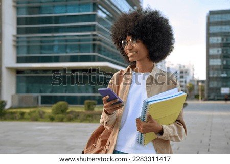 Happy African American student girl with afro haircut enjoy online communication, hold exercise books and backpack, scroll social media app on cell phone device, looking at camera on city street. Royalty-Free Stock Photo #2283311417