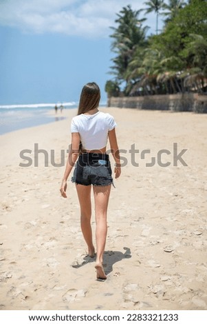 Woman in a white cropped t-shirt walking along a beautiful beach. Back view Mock-up.