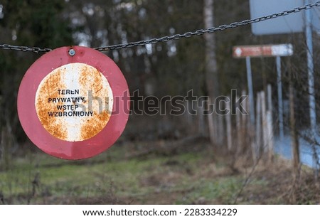 Traffic prohibition sign hung on a chain stretched across the road. A rusty sign that reads " PRIVATE PROPERTY NO ENTER!! " . 