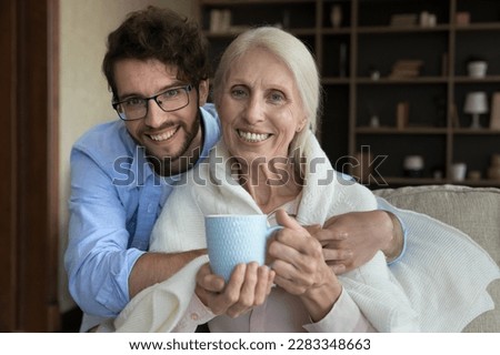 Happy young adult grandson caring for sick elder grandma. Senior lady wrapped in warm plaid suffering from flu, cold, drinking hot tea, enjoying meeting with son, looking at camera, smiling Royalty-Free Stock Photo #2283348663