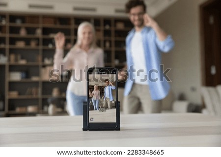 Close up of smartphone screen recording video of young adult son and senior mother posing in home living room, dancing, waving greeting hands hello, standing, smiling, laughing, having fun