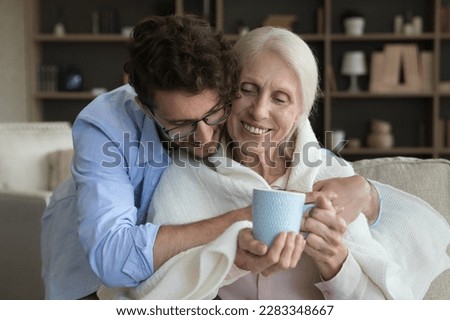 Loving young adult son giving positive sick senior mother help, assist, care, wrapping mom shoulders into plaid, scarf, giving cup of hot drink, hugging, smiling, laughing Royalty-Free Stock Photo #2283348667