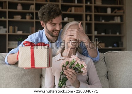 Cheerful adult son guy giving surprise gift to old senior mom, covering woman eyes with hand, holding present box, flowers bouquet, celebrating international women day, birthday, mothers day Royalty-Free Stock Photo #2283348771