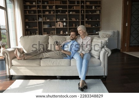 Happy young adult guy resting on sofa near older mother, using smartphone, taking family selfie, talking on video call, laughing, having fun. Senior retired mom hugging joyful grownup son