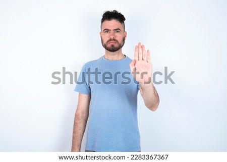 Young caucasian man wearing blue t-shirt over white background shows stop sign prohibition symbol keeps palm forward to camera with strict expression