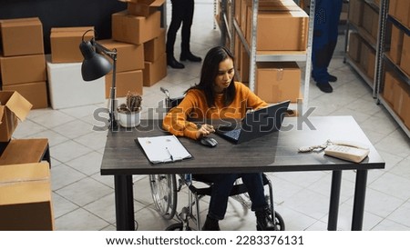 Young employee with impairment working in warehouse, doing desk work for stock logistics and inventory on laptop. Woman wheelchair user doing financial planning in disability friendly space. Royalty-Free Stock Photo #2283376131