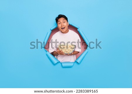 Happy Asian boy standing in the middle of blue background with popcorn bowl in hands up, guy wants to have fun and watch a movie at home, spare time concept, copy space, high quality photo