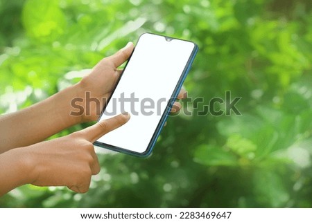 Hand holding smartphone with white blank screen in hand, selecting the application menu in green outdoor environments. Gadget with empty free space for mock up related to environment, healthy, ads