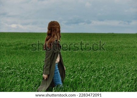 a red-haired woman in a long raincoat stands in a green field in cloudy weather in a strong wind