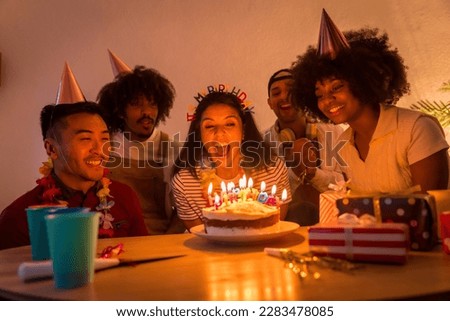 Multi-ethnic group of friends at a birthday party on the sofa at home with a cake and gifts, blowing out the candles with the lights off
