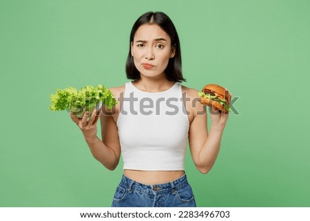 Young minded sad upset woman wear white clothes hold eat burger fresh greens lettuce leaves isolated on plain pastel light green background. Proper nutrition healthy fast food unhealthy choice concept