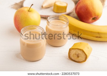 Baby puree with fruits mix, apple, banana infant formula in glass jar on white wooden background. Side view, close up, artificial feeding concept. Royalty-Free Stock Photo #2283537073