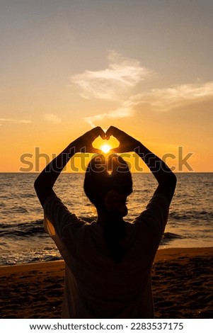 Back view of Travel asian young woman with making heart sign on sea beach at sunset in summer, Thailand.