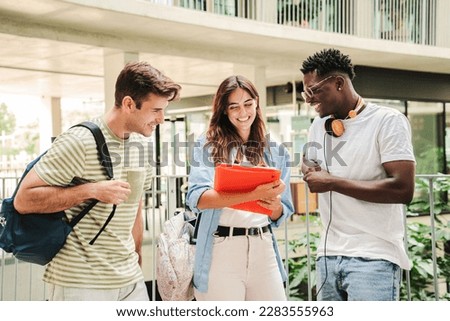 Happy multiethnic group of teenage students talking about the homeworks after the class at university campus. Three young people smiling, having a friendly conversation together standing at the school