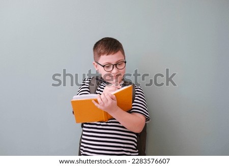 A cute cheerful boy in stripped tee shirt in glasses and is writing a yellow book and with school bag on back on green background.